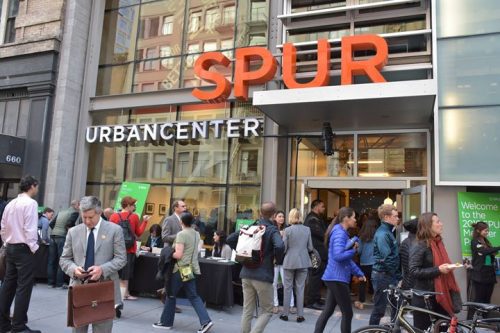 People gather outside the spur urban center during an event, with some attendees checking their phones and others chatting.