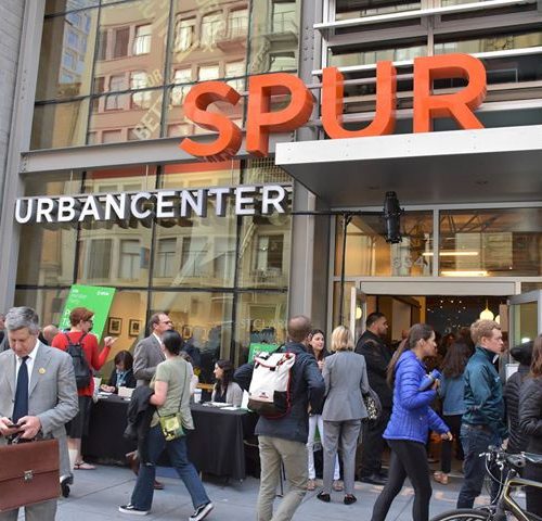 People gather outside the spur urban center during an event, with some attendees checking their phones and others chatting.