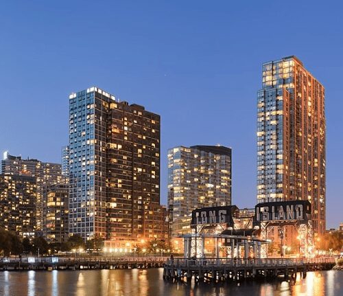 Twilight view of a modern city skyline with illuminated buildings by a waterfront, featuring a 