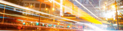 Long-exposure shot of a busy city intersection at night, showcasing vibrant light trails from moving vehicles against illuminated skyscrapers.