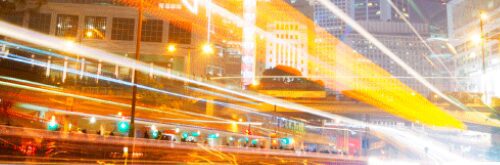 Long-exposure shot of a busy city intersection at night, showcasing vibrant light trails from moving vehicles against illuminated skyscrapers.
