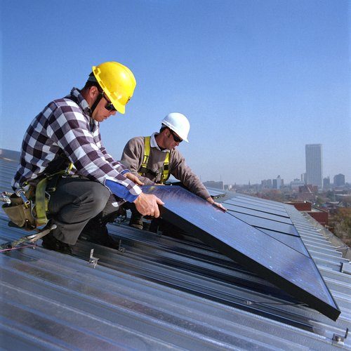 Two workers in helmets install solar panels on a roof under a clear blue sky.