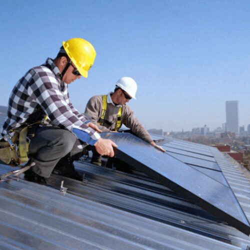 Two workers in hard hats and safety harnesses install solar panels on a metal roof against a city skyline.
