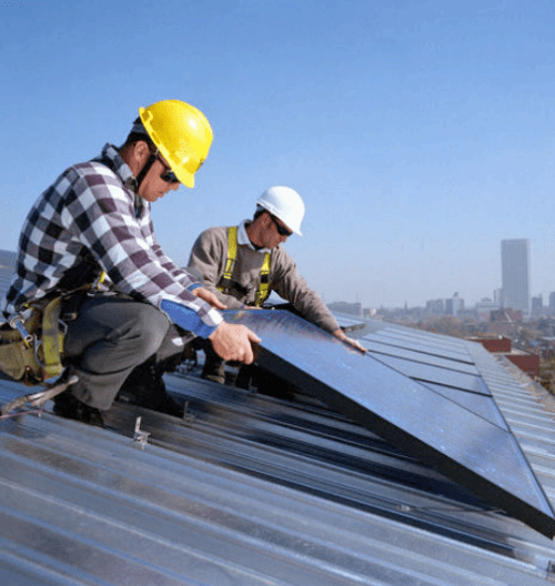 Two workers in hard hats and safety harnesses install solar panels on a metal roof against a city skyline.