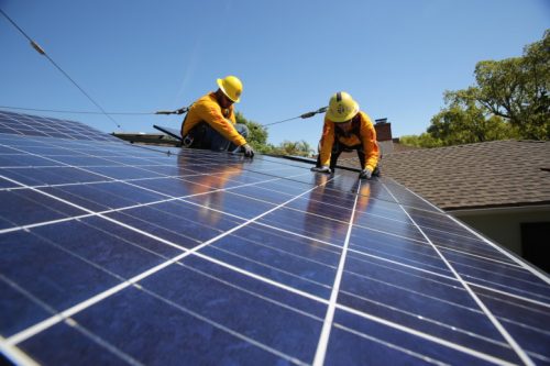 Two workers in yellow hard hats installing solar panels on a residential rooftop on a sunny day.