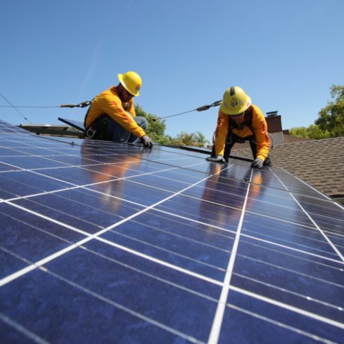 Two workers in yellow hard hats installing solar panels on a residential rooftop on a sunny day.