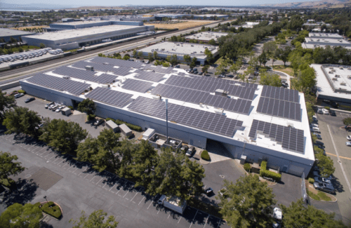 Aerial view of a large industrial building with a roof covered in solar panels, surrounded by a parking lot and green landscape, located next to a highway.