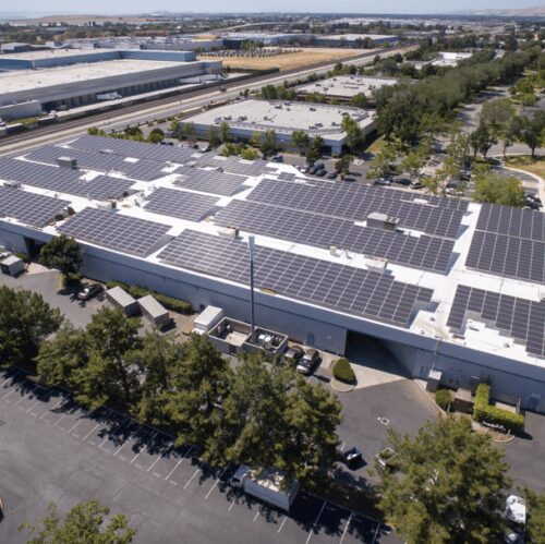 Aerial view of a large industrial building with a roof covered in solar panels, surrounded by a parking lot and green landscape, located next to a highway.