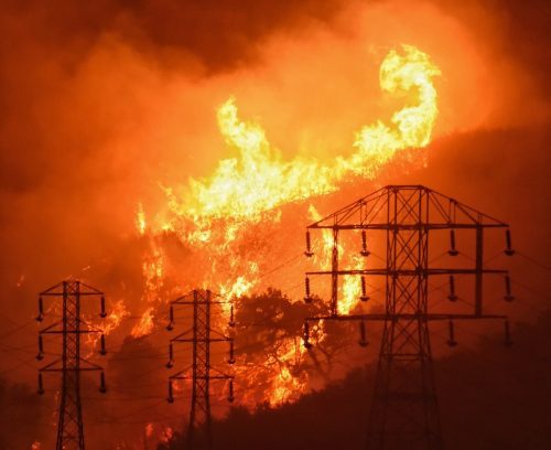 Intense wildfire consuming hillside at night, with flames near power transmission towers.