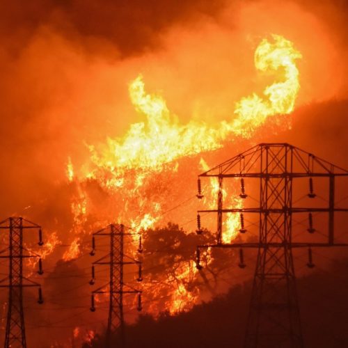 Intense wildfire consuming hillside at night, with flames near power transmission towers.