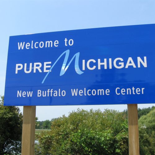 Blue welcome sign reading 'welcome to pure michigan, new buffalo welcome center' against a backdrop of trees and a clear sky.