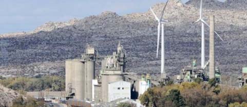Industrial plant with large silos and wind turbines against a mountainous backdrop.