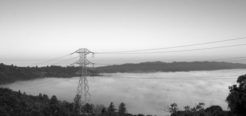 Black and white image of a landscape with a power line tower foreground, overlooking a valley blanketed in thick fog.