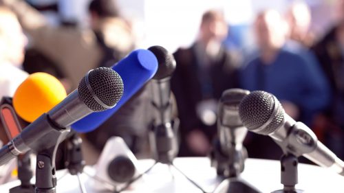 Multiple microphones on a stand with blurred audience in the background, suggesting a press conference or public speaking event.