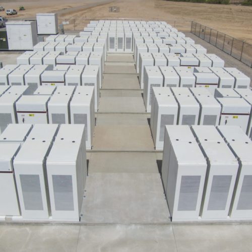 Aerial view of a large battery storage facility with rows of white industrial battery units enclosed by a fence in a desert setting.