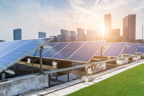 Solar panels on a rooftop with city skyline in the background and sun shining brightly above.