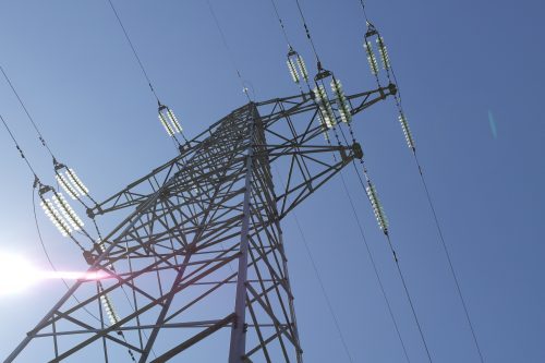 Low-angle view of an electrical transmission tower against a clear blue sky, sunlight glaring slightly on the left.