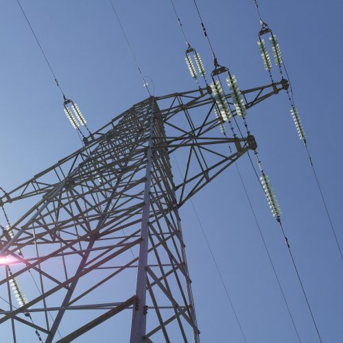 Low-angle view of an electrical transmission tower against a clear blue sky, sunlight glaring slightly on the left.