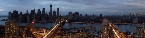 Panoramic view of a city skyline at dusk with prominent high-rise buildings and a lit bridge in the foreground.