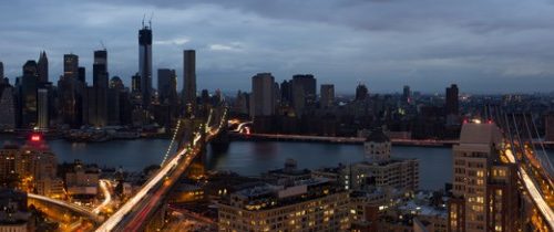 Panoramic view of a city skyline at dusk, featuring tall skyscrapers and illuminated bridges.