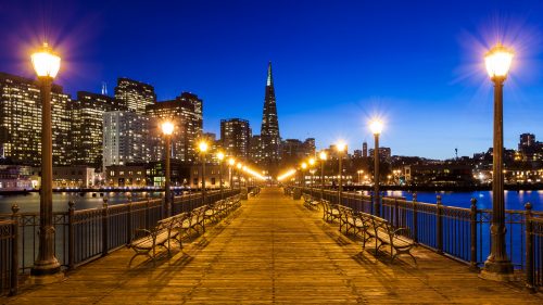 Evening view of a pier leading towards a city skyline with illuminated skyscrapers and street lamps.