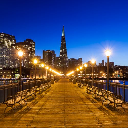 Evening view of a pier leading towards a city skyline with illuminated skyscrapers and street lamps.