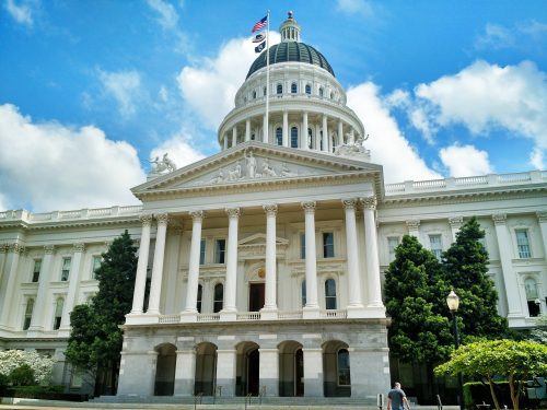 Front view of the california state capitol building with a clear blue sky and a few clouds, featuring neoclassical architecture and a large american flag atop.
