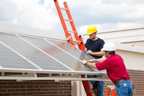 Two technicians, one on a ladder, installing solar panels on a rooftop under a cloudy sky.