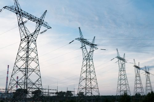 High-voltage transmission towers against a cloudy sky at dusk, with industrial structures in the background.