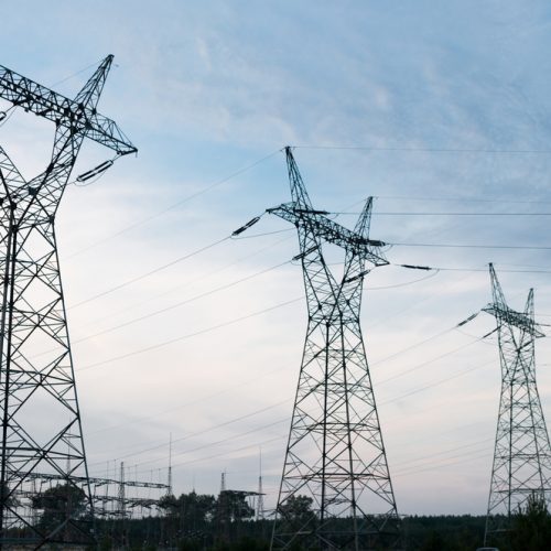 High-voltage transmission towers against a cloudy sky at dusk, with industrial structures in the background.