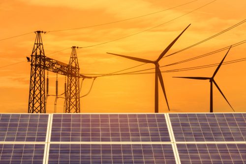 Solar panels in the foreground with wind turbines and an electricity pylon against an orange sky, depicting renewable energy sources.