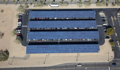 Aerial view of a parking lot covered with solar panels, with cars parked underneath and adjacent roads visible.