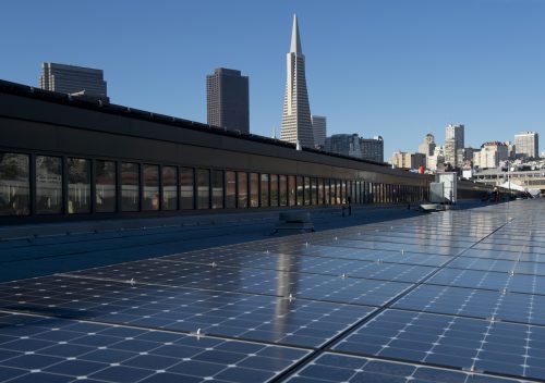 Solar panels on a rooftop with a city skyline including a tall pointed building in the background under a clear blue sky.