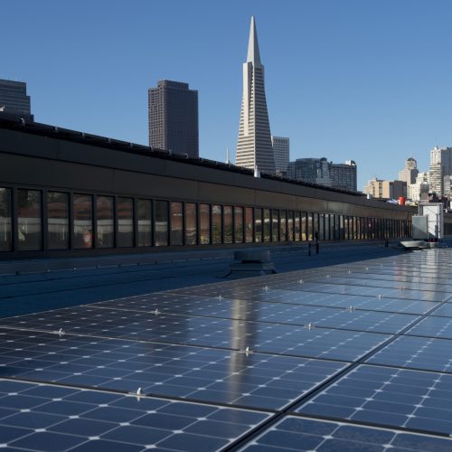 Solar panels on a rooftop with a city skyline including a tall pointed building in the background under a clear blue sky.