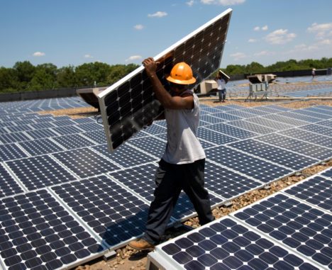 A worker in a hard hat installs a large solar panel at a vast solar farm under a clear blue sky.