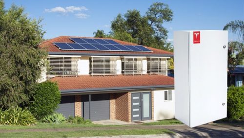 A suburban home with solar panels on the roof and a large tesla powerwall battery next to it on a sunny day.
