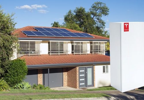 A suburban home with solar panels on the roof and a large tesla powerwall battery next to it on a sunny day.