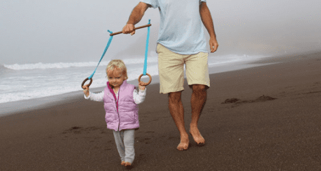 A man and a toddler walking on a sandy beach, with the man holding a harness to support the toddler.