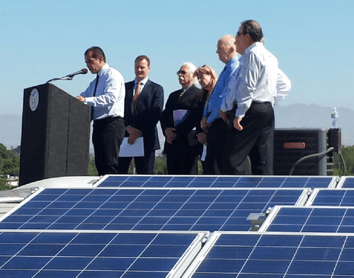A group of officials, including a man speaking at a podium, stands beside solar panels at an outdoor event under a clear sky.
