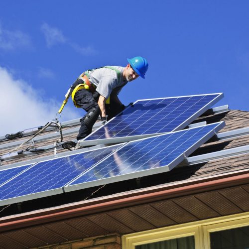 A worker in a blue helmet installs solar panels on a sloped house roof, secured by a safety harness under a clear sky.