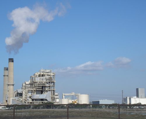 Industrial plant with smokestacks emitting steam against a clear sky, enclosed by a chain-link fence.