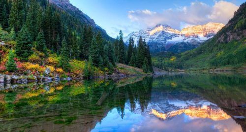 Snow-capped mountains reflected in a calm lake with forested shores under a clear sky at sunrise.