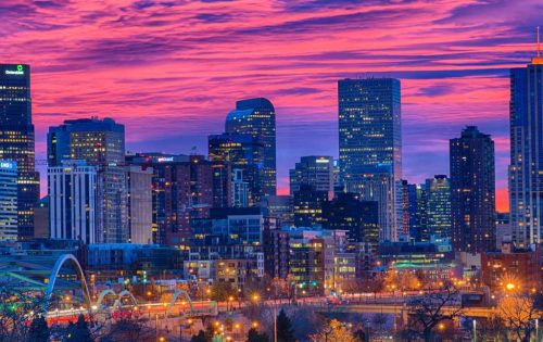 City skyline during sunset with vibrant purple and orange clouds, featuring illuminated buildings and a bridge in the foreground.