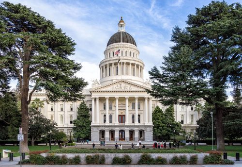 Front view of the california state capitol building in sacramento, framed by trees, with people walking in front.