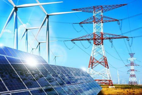 Solar panels and wind turbines in a field under a clear sky, with electricity pylons in the background.