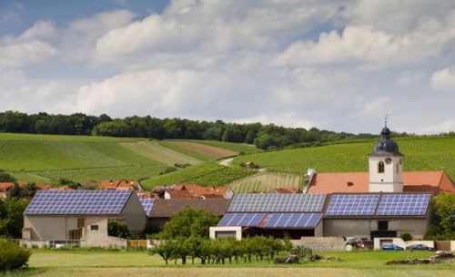 Village houses with solar panels on roofs, a church with a bell tower, and lush green, rolling vineyard hills under a cloudy sky.