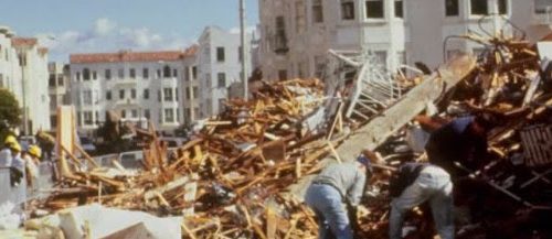 Emergency workers search through rubble on a city street after a severe earthquake, with damaged buildings in the background.