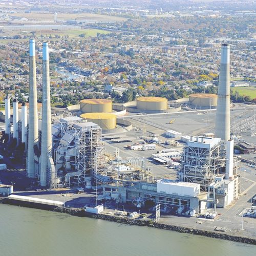 Aerial view of a coastal industrial plant with tall smokestacks and large storage tanks, surrounded by a residential area.