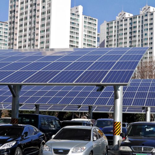 Solar panels installed above parking spaces with parked cars, set against a backdrop of high-rise residential buildings.