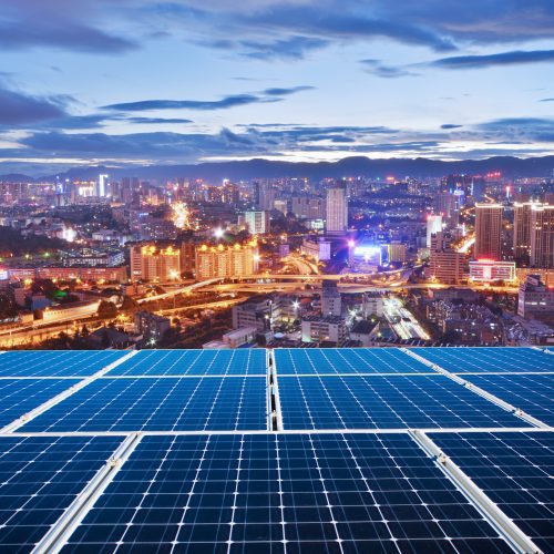 Solar panels in the foreground overlooking a brightly illuminated cityscape during twilight.